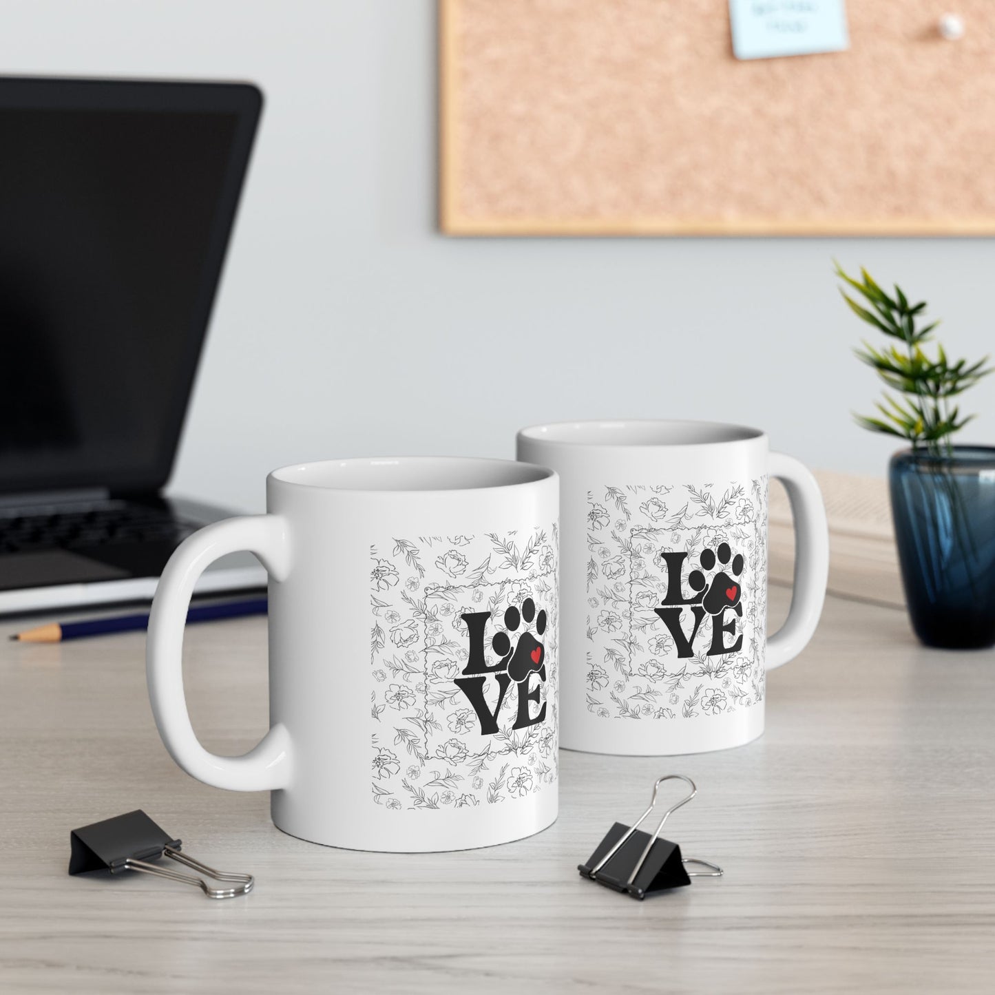 Two white mugs with 'LOVE' and paw print designs on a desk with office supplies.