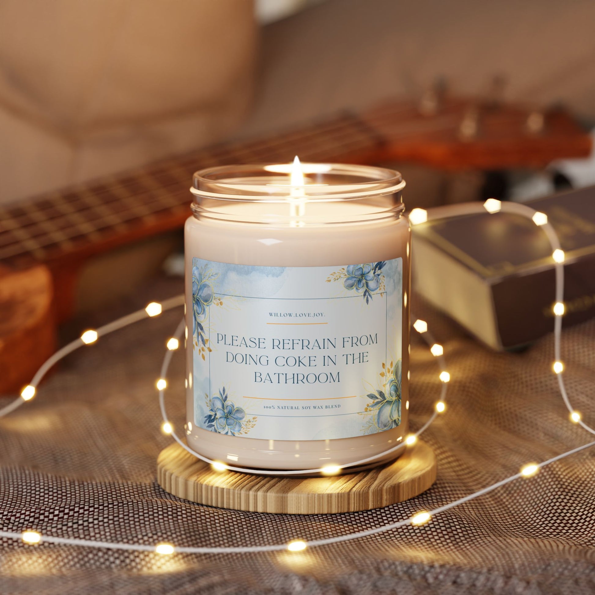 Candle with a humorous label on a wooden coaster surrounded by string lights and a guitar in the background